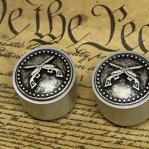May include: Two silver-toned metal knobs with a crossed gun design on the top. The knobs have a black and silver design and are set against a background of a document with text.