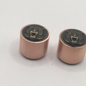 Pu&ograve; includere: Due bottoni cilindrici con finitura metallica color oro rosa. Ogni bottone presenta un inserto circolare sulla superficie superiore che mostra un rilievo rialzato color bronzo antico di una testa di tigre ringhiante con denti visibili.