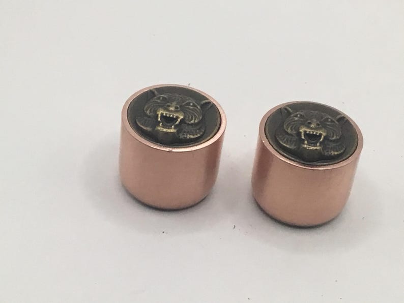 Pu&ograve; includere: Due bottoni cilindrici con finitura metallica color oro rosa. Ogni bottone presenta un inserto circolare sulla superficie superiore che mostra un rilievo rialzato color bronzo antico di una testa di tigre ringhiante con denti visibili.