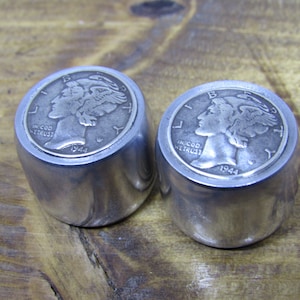 Könnte beinhalten: Zwei silberne Flaschenverschlüsse aus Metall mit einer 1944 Walking Liberty Half Dollar Münze, die oben eingelassen ist. Die Münzen zeigen nach links und haben die Inschriften "IN GOD WE TRUST" und "LIBERTY".