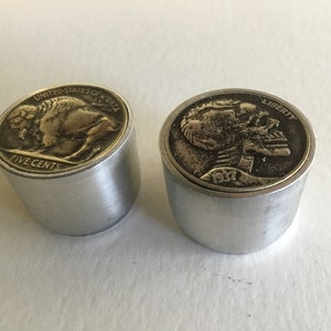 May include: Two silver metal knobs with coin tops. The left knob has a buffalo nickel coin top with the words "United States of America" and "Five Cents". The right knob has a 1937 Liberty nickel coin top with a skull design and the word "Liberty".