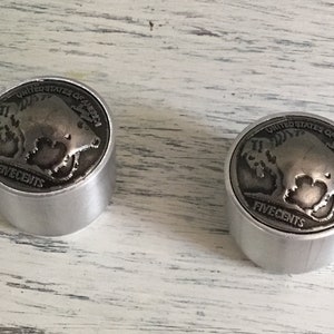 May include: Two silver metal knobs with a buffalo nickel design. The knobs are round and have a smooth surface. The buffalo nickel design is raised and has a detailed image of a buffalo.