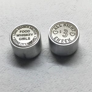 May include: Two silver metal tokens. One token says "Long Branch Saloon * Food Whiskey Girls * Dodge City, Kansas". The other token says "All Night * £3 Check".