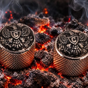 May include: Two cylindrical metal knobs with a textured, diamond-patterned side and a flat top featuring an embossed eagle emblem holding a shield with the letter C in the center. The knobs are positioned against a background of glowing hot coals.