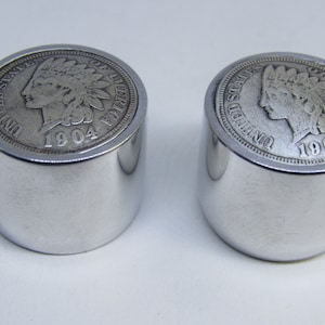 Może przedstawiać: Dwa srebrne metalowe korki do butelek z wzorem 1904 Indian Head penny na górze.