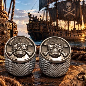 May include: A pair of circular silver-toned cufflinks featuring a raised skull and crossbones design on the face. The sides of the cufflinks have a textured, diamond-patterned finish. The metal has a polished, metallic sheen throughout.