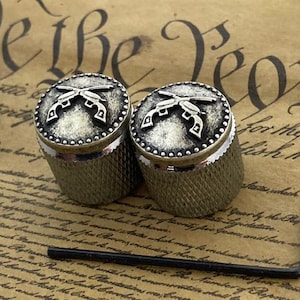 Op de afbeelding: Twee zilveren gitaarknoppen met een ontwerp van gekruiste pistolen. De knoppen hebben een getextureerd oppervlak en een zilveren afwerking.