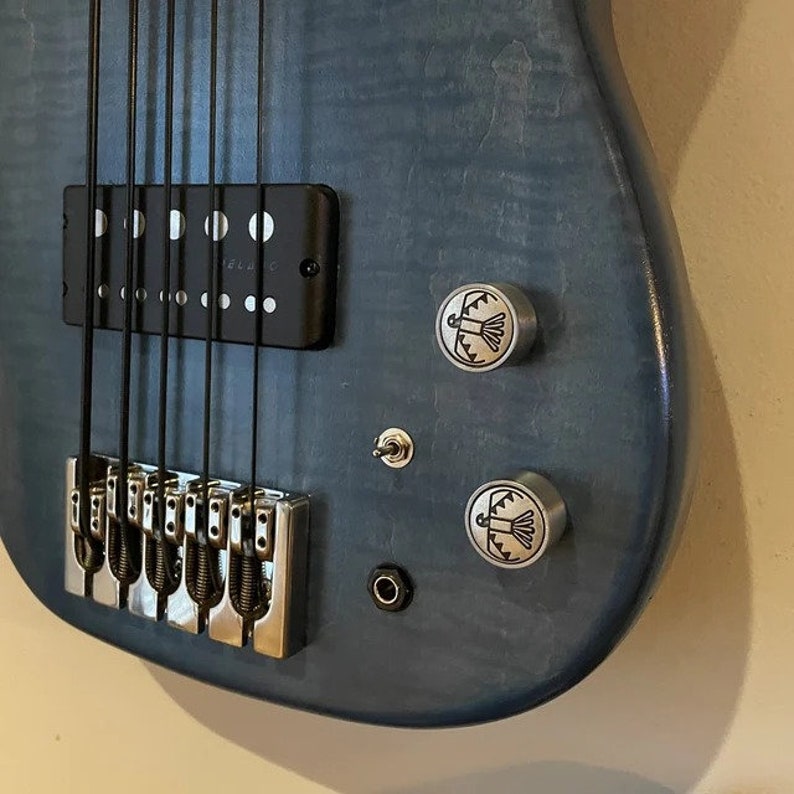 May include: A close-up of a blue bass guitar. The instrument features black strings, a black pickup, and silver hardware. The body has two silver knobs with a bird design and a small switch.