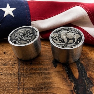 Könnte beinhalten: Zwei zylindrische Metallobjekte mit eingebetteten 1937 Buffalo-Nickel-Münzen. Eine Münze zeigt ein geschnitztes Totenkopfprofil mit dem Text LIBERTY und 1937, die andere einen Büffel mit dem Text UNITED STATES OF AMERICA und FIVE CENTS.