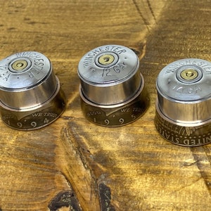 May include: Three bottle stoppers crafted from Winchester 12 GA shotgun shell casings, copper rings, and US coins. The coins are dated 1994, 1995, and 1995. The stoppers are displayed on a wooden surface. The stoppers are a unique decorative item.