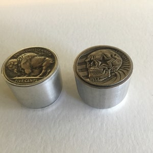 Może przedstawiać: Dwa metalowe przedmioty w kształcie monet z wypukłym wzorem. Lewy przedmiot przedstawia bizona z tekstem "UNITED STATES OF AMERICA" i "FIVE CENTS". Prawy przedmiot przedstawia czaszkę z gwiazdami nad oczami i tekstem "LIBERTY" i "1937".