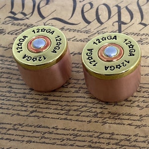 Peut inclure: Deux bouchons de bouteille en cuivre avec des dessus dorés. Les dessus sont gravés avec "12 GA" et une étoile. Les bouchons sont posés sur un morceau de papier avec du texte.