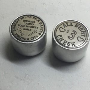 Könnte beinhalten: Zwei silberne Metallmarken. Eine Marke hat den Text "SHADDY MILTS SILK GARTER VIRGINIA CITY" und den Text "BROTHEL CASINO FOOD WHISKEY GIRLS" mit zwei Symbolen. Die andere Marke hat den Text "ALL NIGHT CHECK" mit einem Herzsymbol auf jeder Seite des Textes.