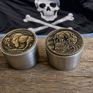 May include: Two cylindrical metal stands topped with coins. The left coin features a buffalo and the text UNITED STATES OF AMERICA FIVE CENTS. The right coin displays a skull wearing star-shaped glasses with the text LIBERTY and the year 1937.