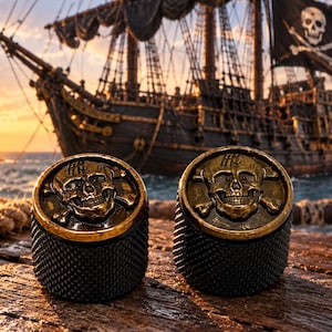 Puede incluir: Dos perillas cilíndricas con un emblema dorado en relieve de una calavera y tibias cruzadas en la superficie superior. Los lados de las perillas tienen un acabado negro texturizado y moleteado. El diseño incluye un pequeño logotipo estilizado.
