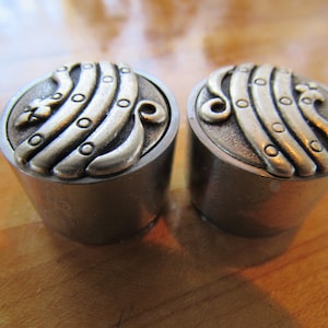 May include: Two silver metal bottle stoppers with a decorative design of a stylized wave pattern in gold metal.