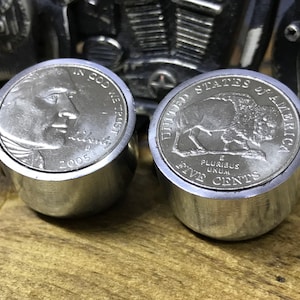 May include: Two silver-colored metal bottle stoppers, each featuring a different design. The left stopper has a 2005 United States nickel coin with the profile of Thomas Jefferson on the front. The right stopper has a 1965 United States nickel coin with the image of a buffalo on the front.