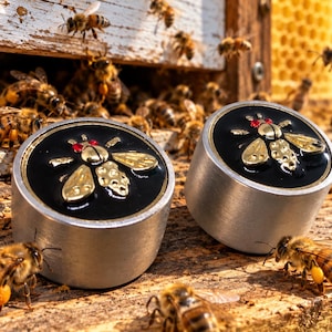 May include: Two cylindrical metal objects with a brushed silver finish and a circular black inlay on top. Each inlay features a raised gold-tone bee emblem with small red accents for eyes, surrounded by numerous live honeybees on a wooden surface.