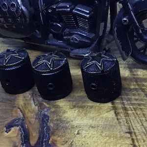 Puede incluir: Cuatro botones de guitarra negros con un diseño en forma de estrella en acabado de latón. Los botones tienen una superficie texturizada y un pequeño agujero en el centro.