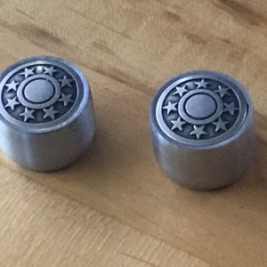 Może przedstawiać: Dwa srebrne metalowe pokrętła z okrągłym wzorem, który zawiera pierścień gwiazd wokół mniejszego koła.