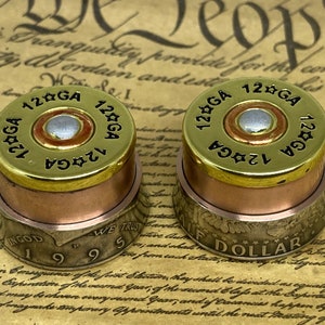 Peut inclure: Deux douilles de cartouches de fusil de chasse de calibre 12 avec des accents en cuivre et en laiton. Les douilles sont empilées sur une pièce d'un dollar argent avec l'année 1995 imprimée dessus. Les douilles sont gravées avec le texte "12 GA" et "12 GA".