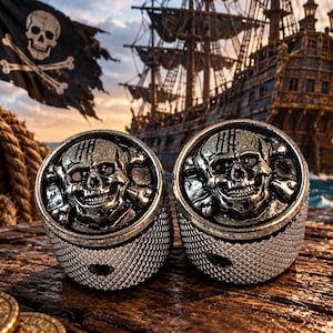 Puede incluir: Dos anillos circulares de metal plateado con un diseño en relieve de calavera y huesos cruzados en la parte frontal. Los lados de los anillos tienen un patrón texturizado de líneas cruzadas. El metal tiene un acabado oscuro y desgastado.
