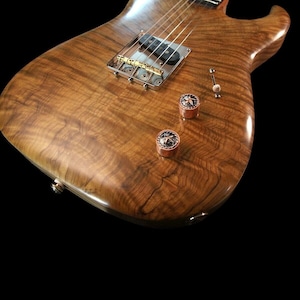 May include: A close-up of a wooden electric guitar with a natural finish. The guitar has a single-coil pickup, two knobs, and a bridge with gold hardware.