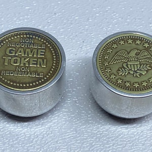 Op de afbeelding: Twee zilveren en messing speltokens. Een token heeft de woorden "Niet onderhandelbaar Speltoken Niet inwisselbaar" erop. De andere token heeft een Amerikaanse zeearend met een banner waarop "USA" staat en een krans van olijftakken.