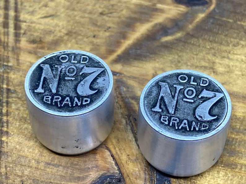 May include: Two cylindrical metal bottle stoppers featuring a raised, dark-toned circular top embossed with the text OLD No. 7 BRAND. The sides of the stoppers are smooth, brushed silver-colored metal with a matte finish and a flat base design.