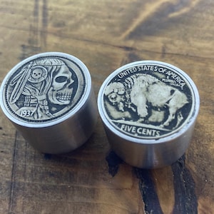 Puede incluir: Dos tapones de botella de metal plateado con diseños grabados. El tapón izquierdo presenta una calavera con un tocado de plumas y el año 1937. El tapón derecho presenta un búfalo con el texto "UNITED STATES OF AMERICA" y "FIVE CENTS".