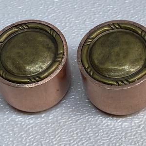 Puede incluir: Dos pomos de cobre y latón con un diseño decorativo. Los pomos son redondos y tienen un centro de latón elevado con un borde de cobre.