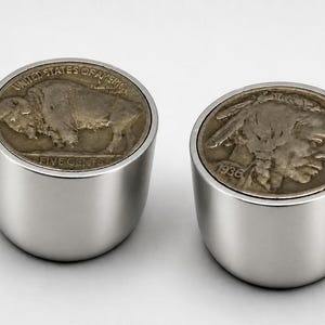 May include: Two cylindrical silver-toned metal objects, each featuring a vintage Buffalo nickel coin embedded in the top. One coin displays a buffalo with the text UNITED STATES OF AMERICA and FIVE CENTS, while the other shows a profile of a person.