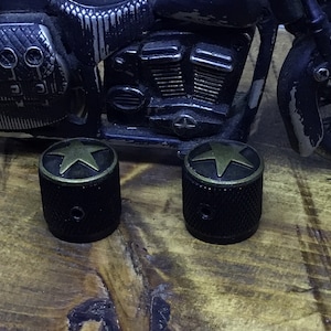May include: Two black metal knobs with a star design and a bronze-colored rim. The knobs are cylindrical and have a textured surface. A toy motorcycle is in the background, resting on a wooden surface.