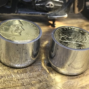 Könnte beinhalten: Zwei silberne Flaschenverschlüsse aus Metall mit goldenen Münzen darauf. Die Münze links zeigt ein Profil eines Mannes mit dem Text "GOD WE TRUST" und "2005". Die Münze rechts zeigt einen Büffel mit dem Text "UNITED STATES OF AMERICA" und "FIVE CENTS".