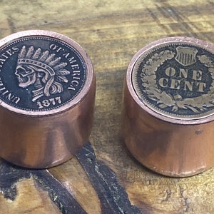 Puede incluir: Dos tapones de botella de cobre, uno con una imagen de una cabeza de nativo americano con una cara de calavera y las palabras "United States of America" y "1877", el otro con una imagen de una corona y las palabras "One Cent".
