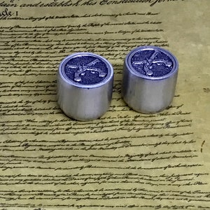 May include: Two silver metal bottle stoppers with a raised design of crossed pistols on the top. The stoppers are sitting on a piece of paper with handwritten text.