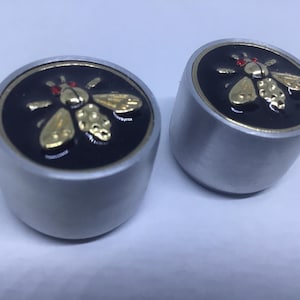 Puede incluir: Dos tapones de botella plateados con fondo negro y un diseño de abeja dorada. La abeja tiene detalles rojos en su cuerpo.