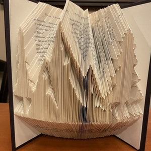 May include: A book art sculpture, also known as a book folding, in the shape of a tree. The book is folded into a 3D shape, with the pages forming the branches of the tree. The book is open, revealing the folded pages.