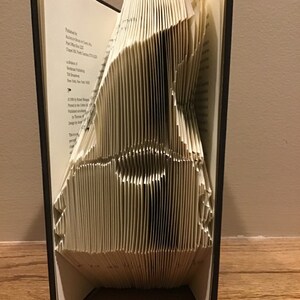 May include: A book with pages folded into a three-dimensional shape, creating a sculpture of a bird. The book is closed and standing upright on a wooden surface.