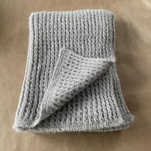 May include: A folded, light gray knitted scarf with a textured, vertical ribbed pattern. The scarf is made of a soft, possibly wool, material. The corner is folded over, revealing a delicate openwork design.