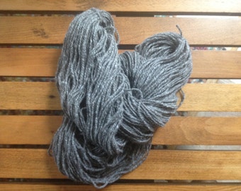 Puede incluir: Un gran ovillo de hilo gris, con sutiles variaciones de tono, descansa sobre una superficie de madera. El hilo parece suave y poco torcido, ideal para proyectos de punto o ganchillo. El color neutro lo hace versátil.
