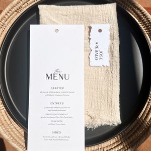 May include: A black plate with a white menu card and a beige napkin, set on a woven wicker charger. The menu lists starters, entrees, and sides. A small tag with the name "JOSE AREBALO" is also present.