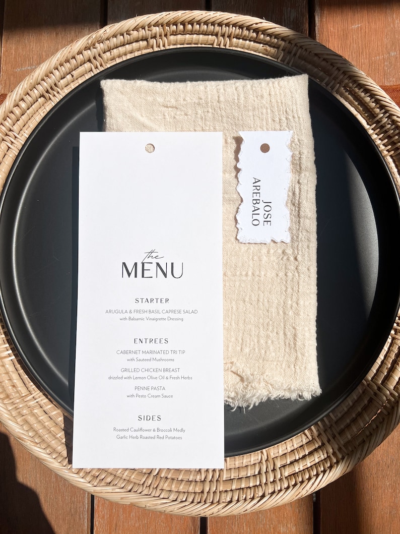 May include: A black plate with a white menu card and a beige napkin, set on a woven wicker charger. The menu lists starters, entrees, and sides. A small tag with the name "JOSE AREBALO" is also present.