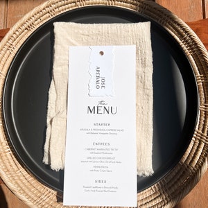 May include: A black plate with a white menu card and a cream napkin, set on a woven placemat. The menu lists starters, entrees, and sides. The menu card has the name "JOSE AREBALO" at the top.