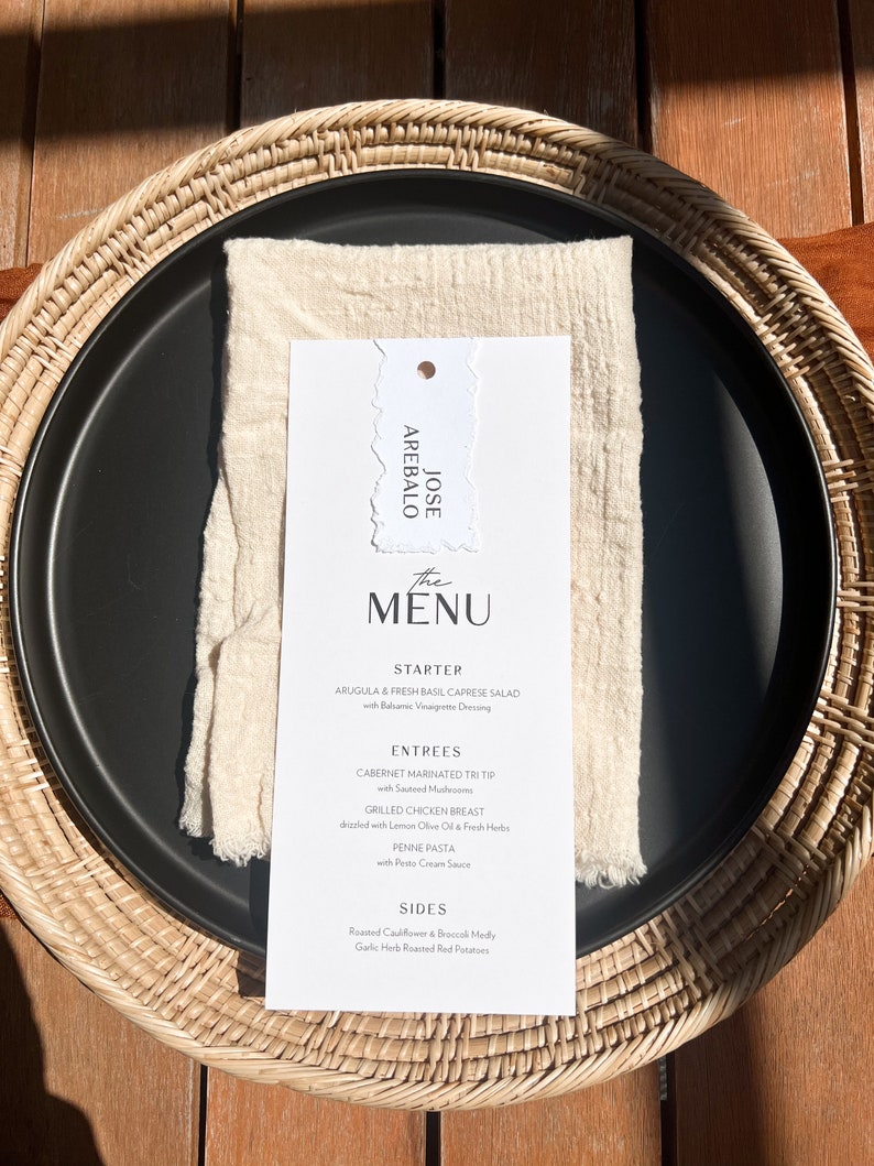 May include: A black plate with a white menu card and a cream napkin, set on a woven placemat. The menu lists starters, entrees, and sides. The menu card has the name "JOSE AREBALO" at the top.