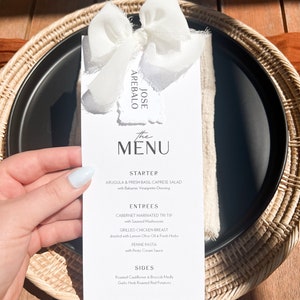 May include: A black plate with a white menu card, tied with a white ribbon. The menu card reads "The Menu" and lists a starter, entrees, and sides. The plate is set on a woven placemat.