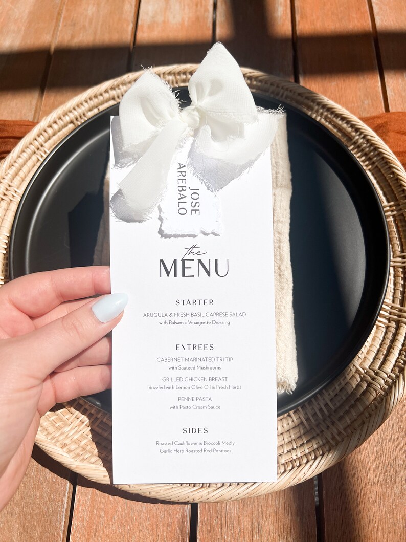 May include: A black plate with a white menu card, tied with a white ribbon. The menu card reads "The Menu" and lists a starter, entrees, and sides. The plate is set on a woven placemat.