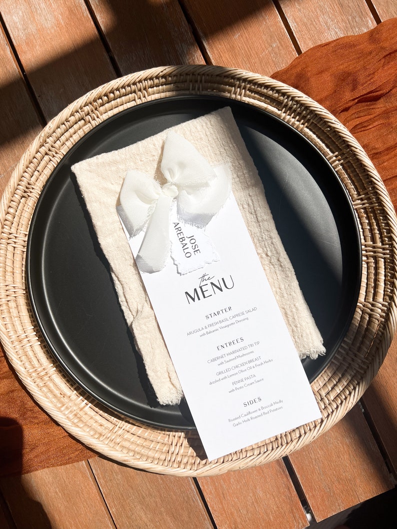 May include: A black plate with a white menu card, a cream-coloured napkin, and a white bow. The menu card has the text "The Menu" and a list of dishes. The plate is on a woven placemat.