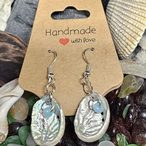 Può includere: Un paio di orecchini d'argento con ciondoli in madreperla di abalone iridescente e perline di vetro blu chiaro. Gli orecchini sono esposti su una carta marrone con il testo "Handmade with love".