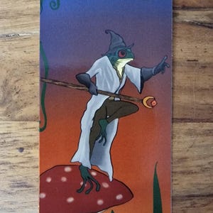 Könnte beinhalten: Ein rechteckiges Lesezeichen mit einer farbenfrohen Illustration eines grünen Frosches, der als Zauberer in einem weißen Gewand und mit spitzem Hut gekleidet ist. Der Frosch steht auf einem großen roten Pilz mit weißen Punkten und hält einen Holzstab.
